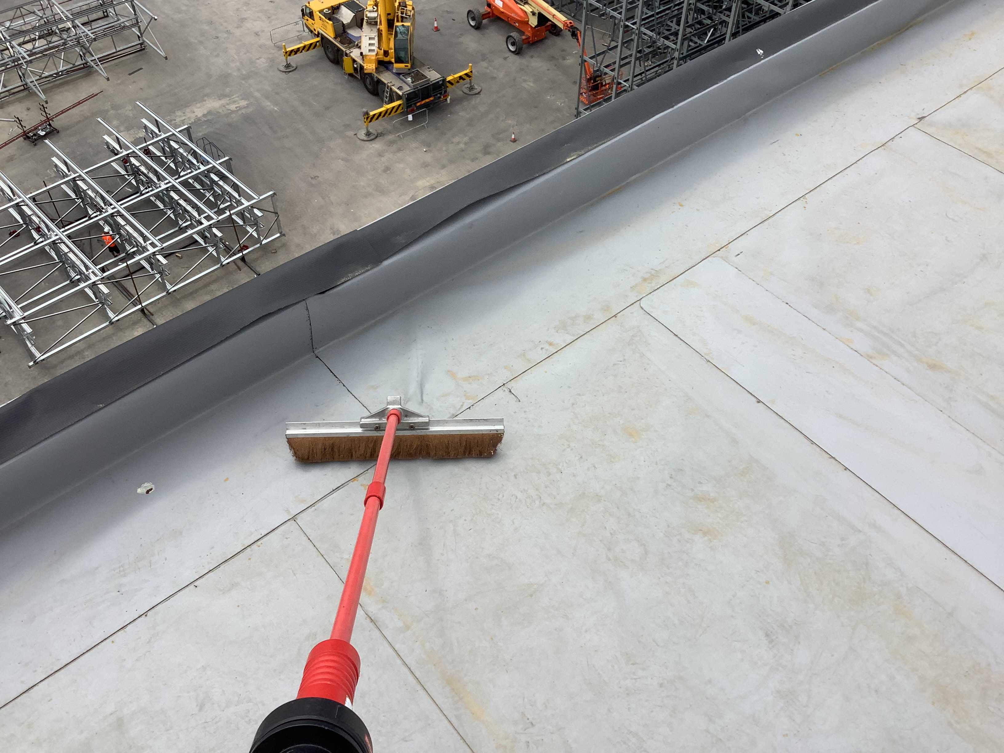 A long-handled brush sweeping a flat roof waterproofing membrane during electronic leak detection preparation, with a construction site visible below.